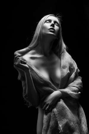 Beautiful young slim busty and leggy naked blonde model girl, sensually covering her nudity with a burlap cloth. Dark background. Conceptual artistic black and white photo.の写真素材