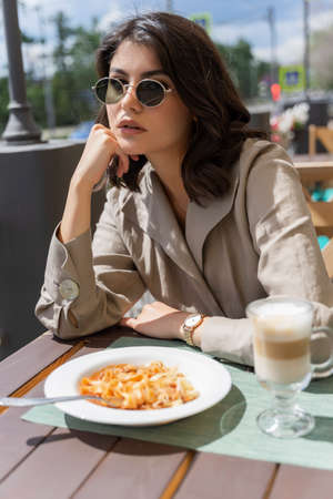 Beautiful smiling brunette girl wearing sunglasses is having lunch in a street cafe, sitting at a...