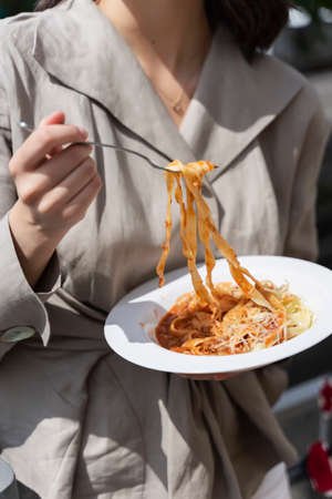 A beautiful smiling brunette girl is having lunch, standing on the street, holding a plate of Italian pasta in her hands. Advertising, commercial design. Copy space.の写真素材