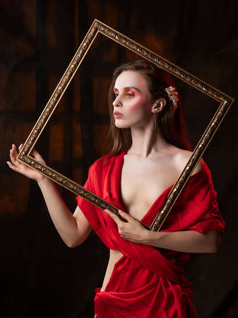 Beautiful girl with trendy makeup, wearing a silk scarf thrown over her shoulders that covers her naked breast, holds picture frame on the background of the shadow from the window.の写真素材