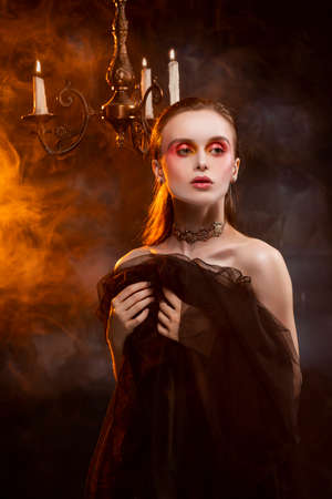A beautiful slim topless girl, wearing earrings and a necklace, and covering her nudity with a black veil, sensually poses in theatrical smoke near the chandelier with lightning candlesの写真素材