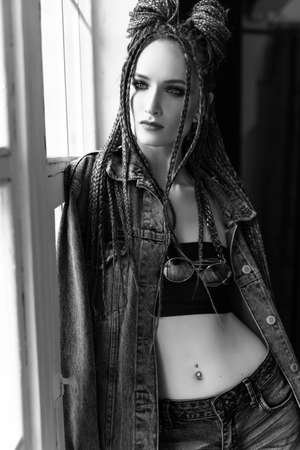 A beautiful young girl with an afro cornrows hairstyle, wearing a casual denim jacket and shorts, relaxes by the window and looks outside thoughtfully. Monochrome, black an white, fashion designの写真素材