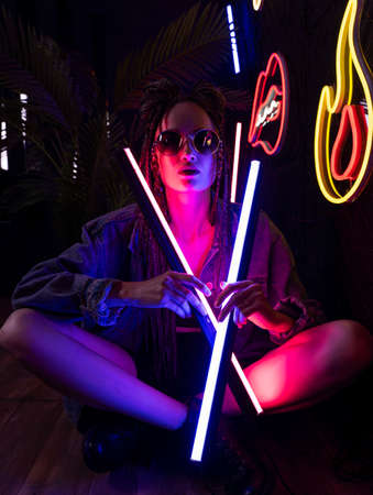 Close up portrait of a beautiful girl with an cornrows, wearing a denim jacket and sunglasses, holds in her hands neon night club lamps. Fashionable, commercial, modern designの写真素材