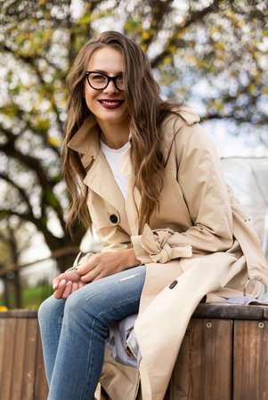 Beautiful smiling girl wearing a light cloak, jeans and a glasses,  joyfully posing, sitting on a bench in an autumn park. Trendy, seasonal, advertising and lifestyle designの写真素材