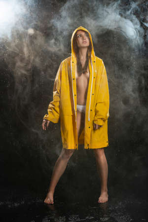 Beautiful topless wet girl wearing white panties and an unbuttoned yellow raincoat posing in scenic smoke under the falling drops of rain on blackの写真素材
