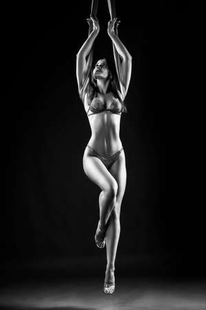 Beautiful brunette girl gymnast wearing lingerie sensually and feminine hanging, posing on canvas. Advertising, commercial and artistic monochrome noir design. Black backgroundの写真素材