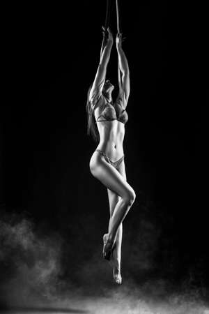 Beautiful brunette girl gymnast wearing lingerie sensually and feminine hanging, posing on canvas. Advertising, commercial and artistic monochrome noir design. Black backgroundの写真素材