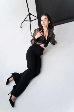 Beautiful Asian girl wearing trousers, a transparent blouse and bra sits on white floor near the gray studio background. Advertising, commercial, fashion designの写真素材