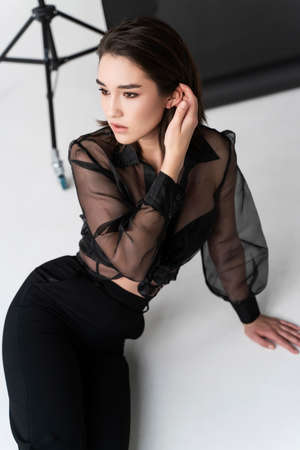 Beautiful Asian girl wearing trousers, a transparent blouse and bra sits on white floor near the gray studio background. Advertising, commercial, fashion designの写真素材
