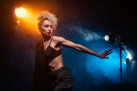 A beautiful blonde girl with an elegant hairstyle and large breasts, wearing a bra, trousers and a blazer, artistically poses in the rays of spotlights in the smoke. Cinematic, art, commercial designの写真素材