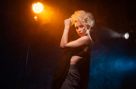 A beautiful blonde girl with an elegant hairstyle and large breasts, wearing a bra, trousers and a blazer, artistically poses in the rays of spotlights in the smoke. Cinematic, art, commercial designの写真素材