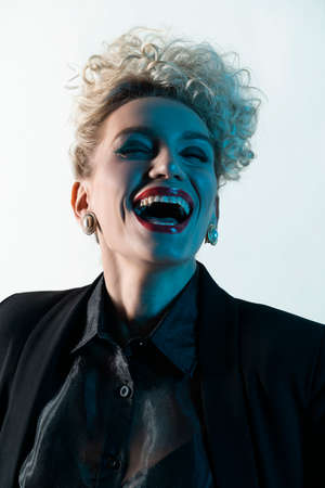 A beautiful blonde girl with an elegant hairstyle and large breasts, wearing a bra, trousers, transparent blouse and a blazer, is laughing in blue light. Trendy, commercial and advertising designの写真素材