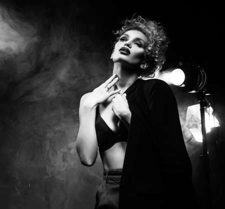 A beautiful blonde girl with an elegant hairstyle and large breasts, wearing a bra, trousers and a blazer, artistically poses in the rays of spotlights in the smoke. Cinematic, art, monochrome designの写真素材