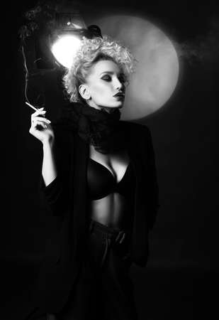A beautiful blonde girl with an elegant hairstyle and large breasts, wearing a bra, trousers, scarf and a blazer, smokes a cigarette near the spotlight. Trendy, monochromeの写真素材
