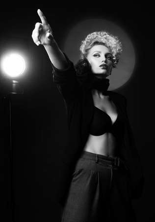 A beautiful blonde girl with an elegant hairstyle and large breasts, wearing a bra, trousers, scarf and a blazer, points forward with her finger. Trendy, monochrome, black and white, noir designの写真素材