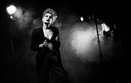 A beautiful blonde girl with an elegant hairstyle and large breasts, wearing a bra, trousers and a blazer, artistically poses in the rays of spotlights in the smoke. Cinematic, art, monochrome designの写真素材