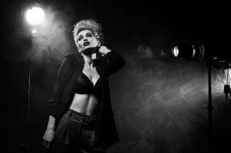 A beautiful blonde girl with an elegant hairstyle and large breasts, wearing a bra, trousers and a blazer, artistically poses in the rays of spotlights in the smoke. Cinematic, art, monochrome designの写真素材