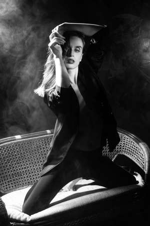 Beautiful braless girl, wearing a unbuttoned blazer, sensually posing dancing on her knees on sofa, on dark background next to a light lamp in the theatrical smoke. Artistic, monochrome, noir designの写真素材