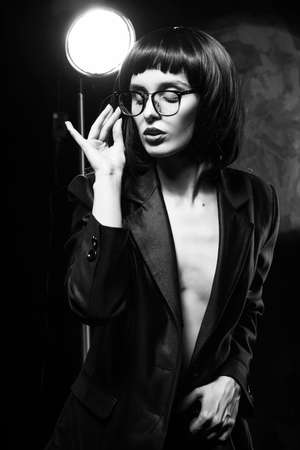 Beautiful braless slim girl with red lips, wearing a unbuttoned black blazer, glasses and brunette wig on a dark background, posing next to the light lamp. Monochrome, trendy, artistic designの写真素材