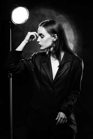Beautiful slim girl model with red lips, wearing a black blazer, touches her face with her hand on a dark background, posing next to a light lamp. Advertising, monochrome, black and white designの写真素材