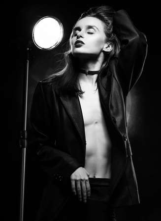 Beautiful braless girl with red lips, wearing an unbuttoned black blazer, touches her hair with her hand on a dark background, posing next to a light lamp. Advertising, trendy, monochrome designの写真素材
