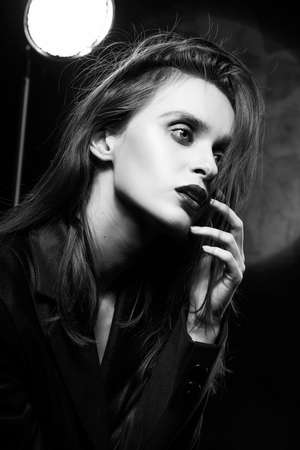 Beautiful braless slim girl, wearing a unbuttoned black blazer, with a slight frenzy and detachment in her eyes touches her face with her hand on a dark background, next to the light lamp. Monochromeの写真素材