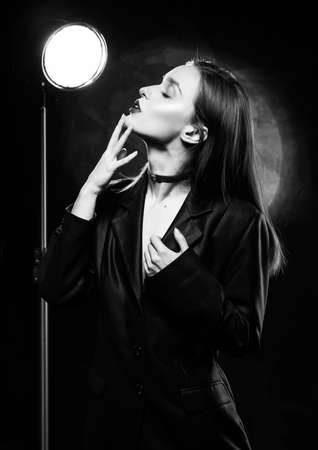 Beautiful slim girl model with red lips, wearing a black blazer, touches her face with her hand on a dark background, posing next to a light lamp. Advertising, monochrome, black and white designの写真素材