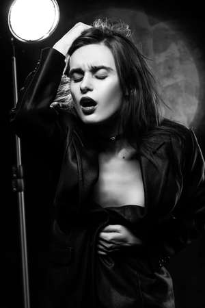 Beautiful slim braless girl, wearing a unbuttoned black blazer, sensually frowns and touches her hair with her hand on a dark background, posing next to a light lamp. Advertising monochrome designの写真素材