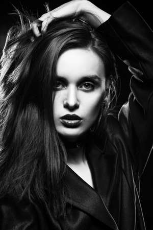 Beautiful slim girl model with red lips, wearing a black blazer, touches her hair with her hand on a dark background, posing next to a light lamp. Advertising, monochrome, black and white designの写真素材