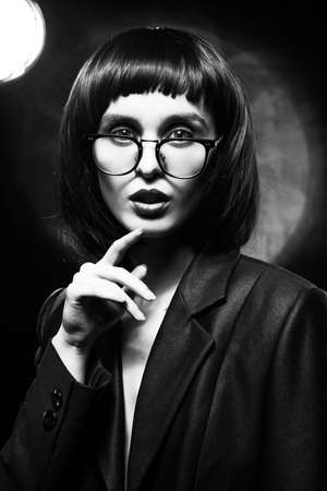 Beautiful braless slim girl with red lips, wearing a unbuttoned black blazer, glasses and brunette wig on a dark background, posing next to the light lamp. Monochrome, trendy, artistic designの写真素材