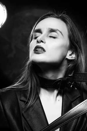 Braless girl with red lips, wearing a unbuttoned blazer and a leather belt tightened around the neck, which she pulls with her hands,  on the dark background, next to a light lamp. Monochrome designの写真素材