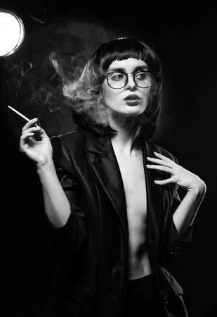 Beautiful braless slim girl with red lips, wearing a unbuttoned black blazer, glasses and brunette wig sensually smokes un a dark background next to the light lamp. Artistic monochrome noir designの写真素材