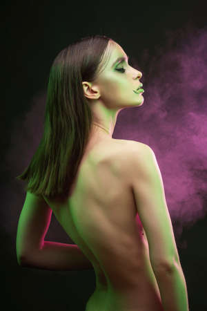 A beautiful slim topless girl with avant-garde makeup sensually poses covering her breast with her hands, in smoke on a black background.  Artistic, fashionable, advertising designの写真素材