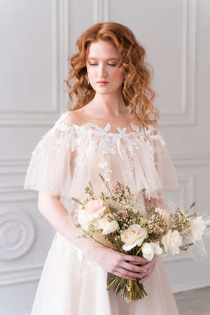 Beautiful natural redhead girl bride, with nude makeup, wearing a white dress, holds a wedding bouquet in her hands, standing in a light interiorの写真素材