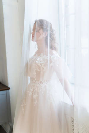 Beautiful natural redhead girl bride, with nude makeup, wearing a white dress, stands at the window behind a transparent curtainの写真素材