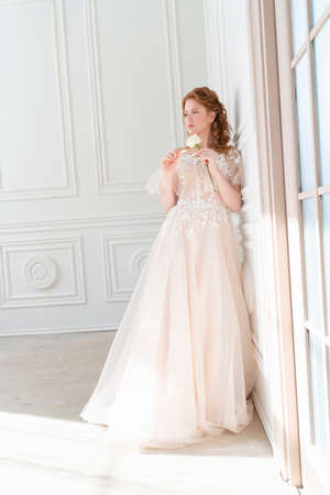 Beautiful natural redhead girl bride, with nude makeup, wearing a white dress,  stands at the white wall, holding a white rose in her hands in light interiorの写真素材