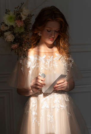 Beautiful natural redhead girl bride, with nude makeup, wearing a white dress, poses in the shadow, illuminated by a narrow beam of light from the window, opens the letter envelopeの写真素材