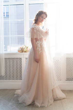 Beautiful natural redhead girl bride, with nude makeup, wearing a white dress, stands at the window next to a transparent curtainの写真素材