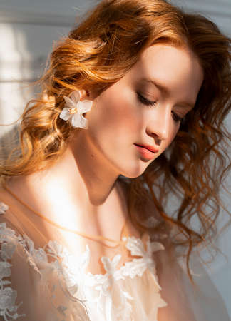 Beautiful natural redhead girl bride, with nude makeup, wearing a white dress, poses in the shadow, illuminated by a narrow beam of light from the window. Close up portraitの写真素材