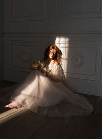 Beautiful natural redhead girl bride, with nude makeup, wearing a white dress, holds a wedding bouquet and sits on the floor in the shadow, illuminated by a narrow beam of light from the windowの写真素材