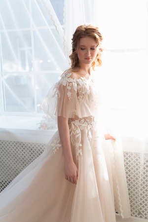 Beautiful natural redhead girl bride, with nude makeup, wearing a white dress, stands at the window next to a transparent curtainの写真素材