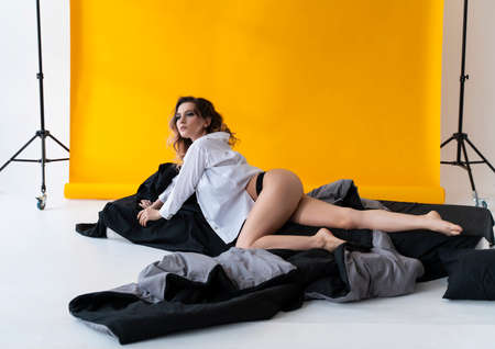 Beautiful busty girl wearing trendy lingerie and unbuttoned shirt, sensually relaxes in the bed near the yellow background. Fashionable, advertising, lifestyle and commercial designの写真素材