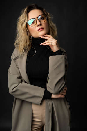 Beautiful girl wearing a casual trendy trousers, turtleneck glasses and raincoat posing on a dark background. Fashionable, advertising, lifestyle and commercial designの写真素材