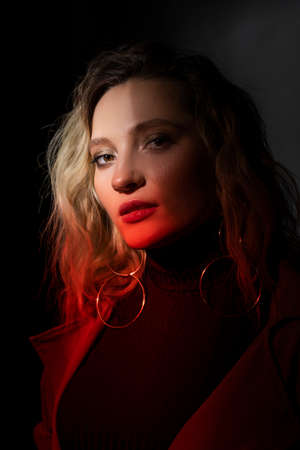 Beautiful girl wearing a casual trendy trousers, turtleneck glasses and raincoat posing on a dark background in the shadows and red light. Fashionable, advertising, lifestyle and commercial designの写真素材