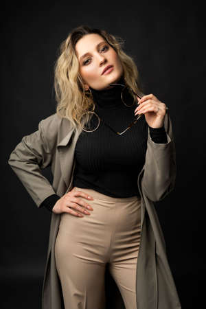 Beautiful girl wearing a casual trendy trousers, turtleneck glasses and raincoat posing on a dark background. Fashionable, advertising, lifestyle and commercial designの写真素材