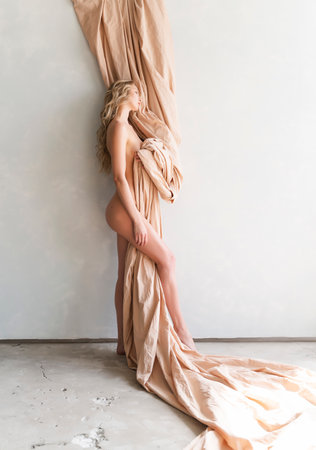 A beautiful blonde curly leggy big breasted naked girl sensually poses sitting on the floor at the wall, covering herself with a large piece of beige cloth. Copy space. Artistic design.の写真素材