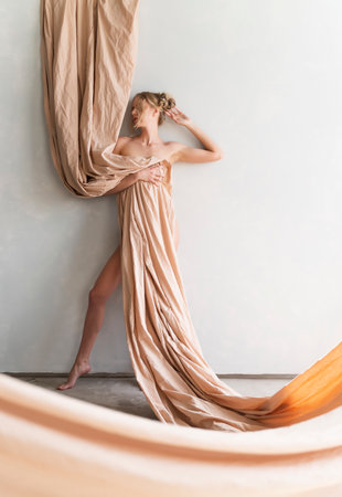 A beautiful blonde curly leggy big breasted naked girl sensually poses sitting on the floor at the wall, covering herself with a large piece of beige cloth. Copy space. Artistic design.の写真素材