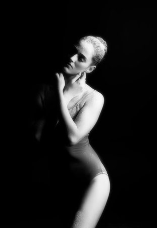 A beautiful slender ballet dancer girl wearing a bodysuit, sensually poses among the flying flour which covers her body, on a black background. Artistic, commercial, monochrome design.の写真素材