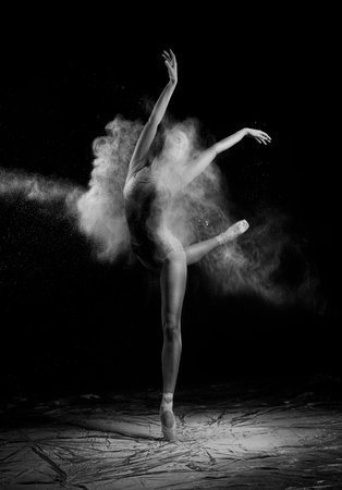 A beautiful slender ballet dancer girl wearing a bodysuit, sensually poses among the flying flour which covers her body, on a black background. Artistic, commercial, monochrome design.の写真素材