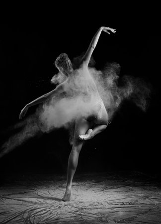 A beautiful slender ballet dancer girl wearing a bodysuit, sensually poses among the flying flour which covers her body, on a black background. Artistic, commercial, monochrome design.の写真素材